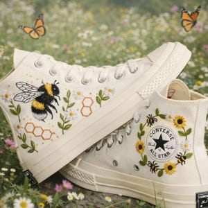 Puede incluir: Zapatillas altas color crema con diseños bordados de abejas y flores. Una zapatilla presenta una abeja grande con panales, margaritas y hojas. La otra zapatilla tiene una corona de girasoles y abejas alrededor del logotipo de Converse. Mariposas revoloteando en el fondo.