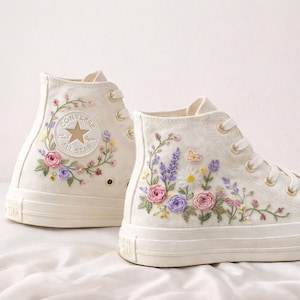 Puede incluir: Zapatillas altas color crema con diseños florales bordados. Las zapatillas presentan un logotipo de Converse All Star y están adornadas con flores rosas, moradas y amarillas y hojas verdes. Cordones blancos y suela crema completan el look.