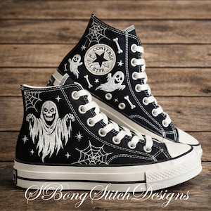 May include: Black high-top sneakers with white embroidered Halloween designs. The sneakers feature a ghost, spiderwebs, bones, and stars. The Converse All Star logo is visible on the side. The text "S. Bong Stitch Designs" is at the bottom.