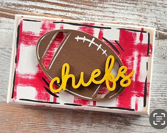 May include: Wooden sign with a red and white checkered background. A brown football cutout with a yellow "chiefs" word cutout is on top of the sign.
