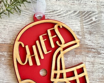 Op de afbeelding: Rode en bruine houten kerstversiering in de vorm van een voetbalhelm met het woord "CHIEFS" uitgesneden aan de voorkant.