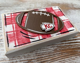 May include: A wooden block with a red and white checkered background. A brown football with white stitching and a white arrow with "KC" in red is on top of the block.