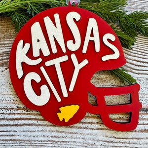 May include: A red wooden ornament shaped like a football helmet, with the words "KANSAS CITY" in white. A yellow arrowhead is on the side. The ornament hangs from a white ribbon and is displayed on a wooden surface with greenery.