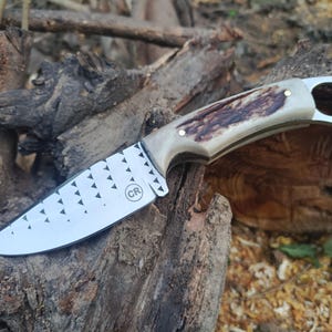 May include: A fixed-blade knife with a silver-colored steel blade featuring a pattern of small triangles and a circular CR logo. The handle is crafted from textured brown and cream-colored antler material, secured with two metal pins and a pommel ring.