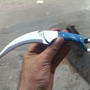 May include: A curved karambit knife with a polished silver blade and a handle featuring blue and white marbled resin. The knife has a finger ring and a small circular marking on the blade. The knife is held in a hand.