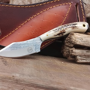 May include: A fixed-blade knife with a textured metal blade featuring a CR logo, a handle made of natural antler with brass pins, and a brown leather sheath with a diamond-patterned embossed design and a circular CR logo stamp.