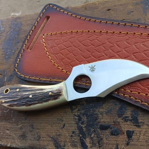 Puede incluir: Un pequeño cuchillo plateado con un mango de asta de ciervo marrón y blanco. El cuchillo tiene un agujero para el dedo en la hoja y un diseño en forma de estrella en la hoja. El cuchillo está en una funda de cuero marrón.