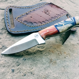 May include: A handcrafted knife with a stainless steel blade and a handle featuring red, white, and blue swirls. The knife rests on a stone surface next to a blue leather sheath with gold stitching. The knife has the initials "CR" on the blade.