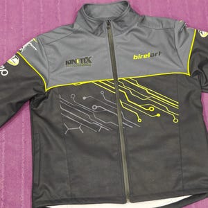 Peut inclure: Veste zippée noire et grise avec des détails jaunes. La veste porte les inscriptions "Kinetix" et "birelart". Un motif de circuit imprimé jaune est présent sur le bas du devant. La veste a un col montant.