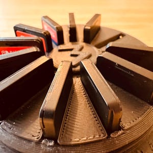 Puede incluir: Un soporte de cartuchos de juego de plástico negro con un diseño circular. El soporte tiene ranuras para varios cartuchos y un centro con un logotipo. Los cartuchos están dispuestos en forma radial alrededor del centro.