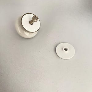 Könnte beinhalten: Ein weißer Keramik-Schrankknopf mit goldfarbenem Metallrand und silberfarbener Schraube. Eine separate weiße Scheibe mit einem zentralen Loch ist ebenfalls vorhanden.