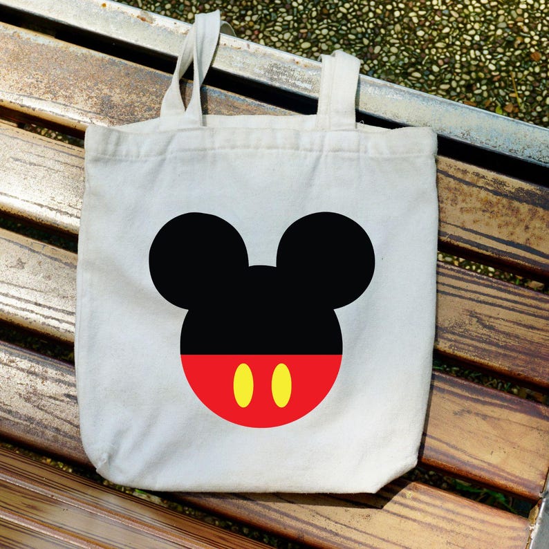 Puede incluir: Una bolsa de mano de lona blanca con un gr&aacute;fico grande de la cara de Mickey Mouse. El dise&ntilde;o incluye orejas negras, una cara negra, una cara inferior roja y dos &oacute;valos amarillos. La bolsa tiene dos asas blancas.
