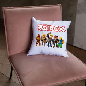 Peut inclure: Coussin carr&eacute; blanc avec le mot "ROBLOX" en lettres rouges. Le coussin pr&eacute;sente un graphisme de cinq personnages de dessins anim&eacute;s dans diverses tenues. Le coussin est pos&eacute; sur une chaise rose.
