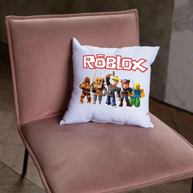 Peut inclure: Coussin carr&eacute; blanc avec le mot "ROBLOX" en lettres rouges. Le coussin pr&eacute;sente un graphisme de cinq personnages de dessins anim&eacute;s dans diverses tenues. Le coussin est pos&eacute; sur une chaise rose.