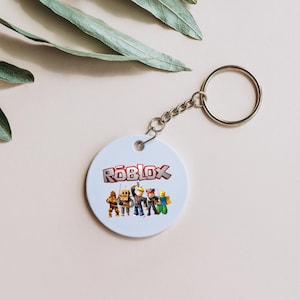 Peut inclure: Un porte-cl&eacute;s rond blanc avec le logo Roblox rouge et blanc, et des personnages de dessins anim&eacute;s en dessous. Il est dot&eacute; d'une cha&icirc;ne et d'un anneau argent&eacute;s.