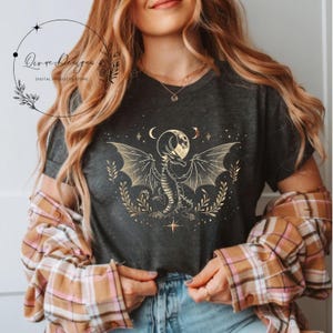 May include: A dark gray t-shirt with a gold graphic of a dragon with wings spread wide. The dragon is standing in front of a crescent moon and stars. The text "Qive Designs" is in the top left corner of the image.
