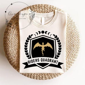 May include: A white t-shirt with a black and gold design. The design features a shield with a dragon inside, a laurel wreath, and the text "Riders Quadrant".