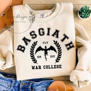 May include: A cream-colored sweatshirt with a black graphic design. The graphic features a dragon with the text "Basgiath", "Fly or Die", and "War College".