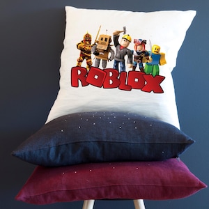 Peut inclure: Une pile de trois coussins d&eacute;coratifs. Le coussin sup&eacute;rieur est blanc avec un graphisme Roblox color&eacute; et le mot "ROBLOX" en rouge. Le coussin du milieu est bleu marine et le coussin inf&eacute;rieur est bordeaux.