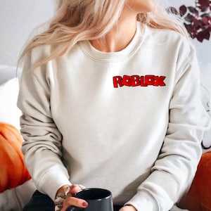 Peut inclure: Sweat-shirt cr&egrave;me avec le mot "ROBLOX" en rouge, bord&eacute; de noir, sur la poitrine. Le sweat-shirt a un col rond et des manches longues. L'image montre une personne portant le sweat-shirt.