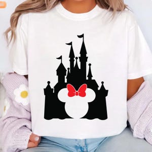 Puede incluir: Camiseta blanca con una silueta negra de un castillo, que incorpora la cabeza de Minnie Mouse y un lazo rojo. El dise&ntilde;o est&aacute; centrado, ofreciendo una imagen clara y reconocible.