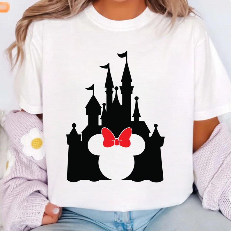 Puede incluir: Camiseta blanca con una silueta negra de un castillo, que incorpora la cabeza de Minnie Mouse y un lazo rojo. El dise&ntilde;o est&aacute; centrado, ofreciendo una imagen clara y reconocible.