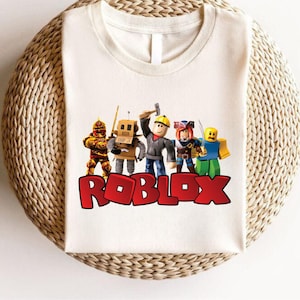 Peut inclure: T-shirt blanc avec un motif de personnages Roblox. Le mot "ROBLOX" est imprim&eacute; en rouge en bas. Le t-shirt a un col rond et des manches courtes. Le t-shirt est pr&eacute;sent&eacute; sur une surface tiss&eacute;e.