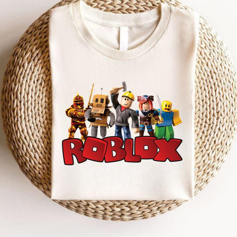 Peut inclure: T-shirt blanc avec un motif de personnages Roblox. Le mot "ROBLOX" est imprim&eacute; en rouge en bas. Le t-shirt a un col rond et des manches courtes. Le t-shirt est pr&eacute;sent&eacute; sur une surface tiss&eacute;e.