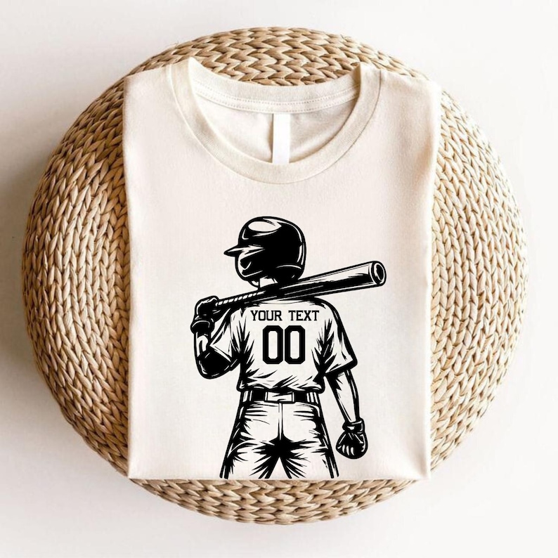 May include: Cream-colored t-shirt featuring a black graphic of a baseball player holding a bat over their shoulder. The shirt has the text "YOUR TEXT" above the number "00" on the back. The player is wearing a helmet and gloves.
