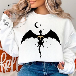 May include: White sweatshirt with a black silhouette of a dragon flying in front of a crescent moon and stars.