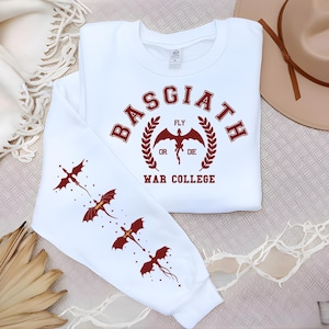 May include: White sweatshirt with the text "BASGIATH WAR COLLEGE" in maroon. The design includes a dragon emblem and the words "FLY OR DIE." The left sleeve features three maroon dragon illustrations. A casual, long-sleeved top.