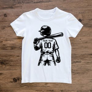 May include: White t-shirt featuring a black and white illustration of a baseball player holding a bat. The player wears a helmet and the shirt displays "YOUR TEXT" above the number "00". The design is a graphic representation of a baseball theme.
