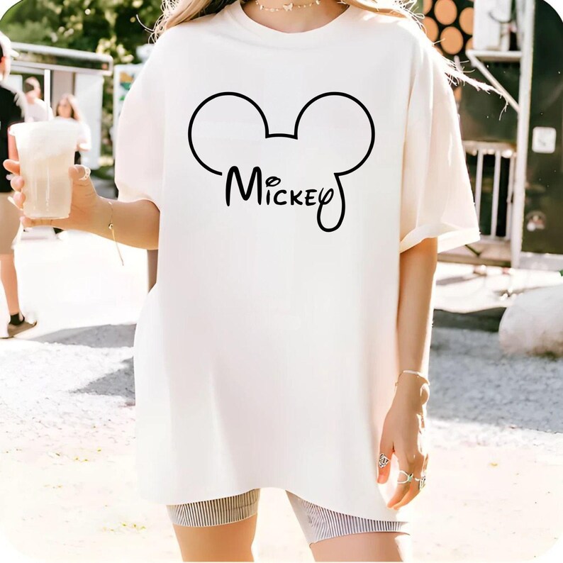 Puede incluir: Camiseta crema de gran tama&ntilde;o con un contorno negro de Mickey Mouse y el nombre "Mickey" en cursiva. La camiseta es de estilo casual y c&oacute;modo.