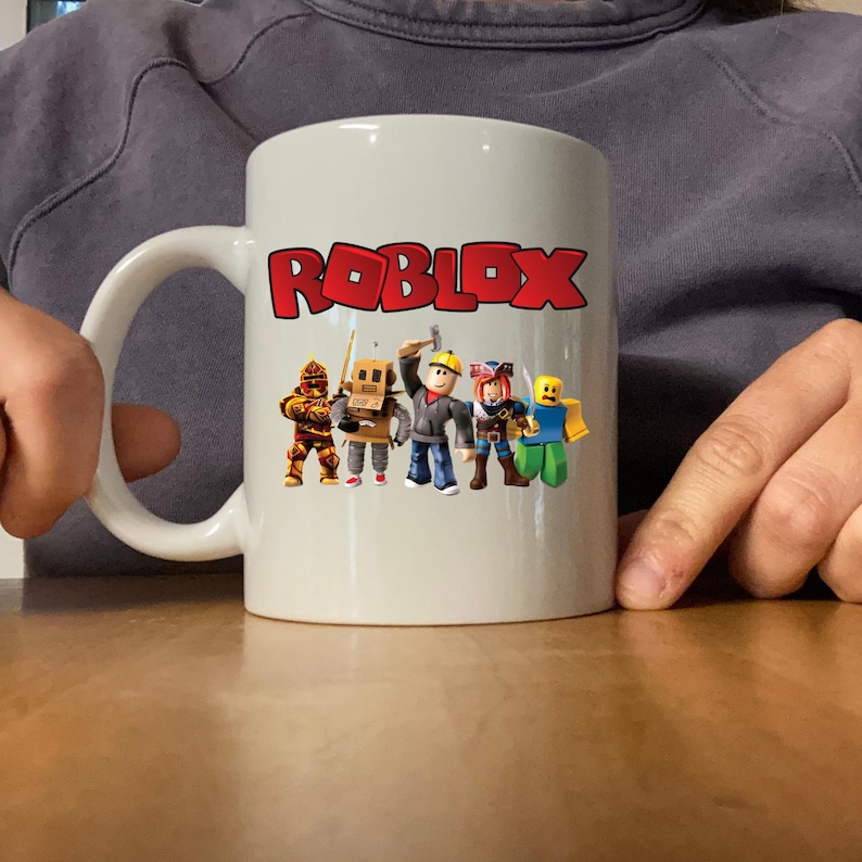 Peut inclure: Mug en c&eacute;ramique blanche avec le mot "ROBLOX" en lettres rouges et un graphisme de personnages de dessins anim&eacute;s. La tasse a une anse blanche et est pos&eacute;e sur une surface en bois.