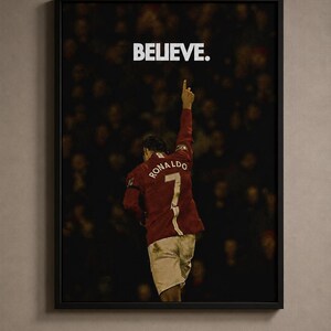 May include: A framed wall poster featuring a soccer player from behind, wearing a red jersey with the name RONALDO and the number 7 on the back. The player has one arm raised toward the sky. Above the player, the word BELIEVE is printed in white.