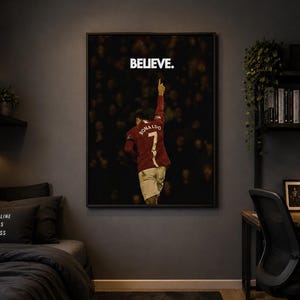 May include: A framed rectangular wall art print featuring a soccer player wearing a red jersey with the name RONALDO and the number 7 on the back, pointing a finger upward. The word BELIEVE. is printed in white capital letters above the player.