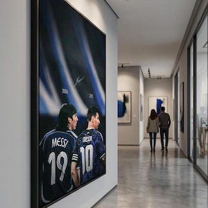 May include: A rectangular framed wall art piece featuring two depictions of Lionel Messi from behind, wearing jerseys with the numbers 19 and 10. The years 2006 and 2026 are printed above the figures, with a signature centred at the top.