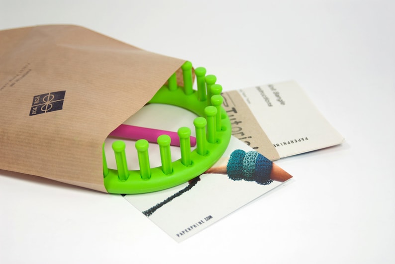 May include: A green plastic knitting loom with a pink knitting needle, a brown paper envelope, and a printed instruction sheet with the text "Tutorials" and "Paperpine.com".