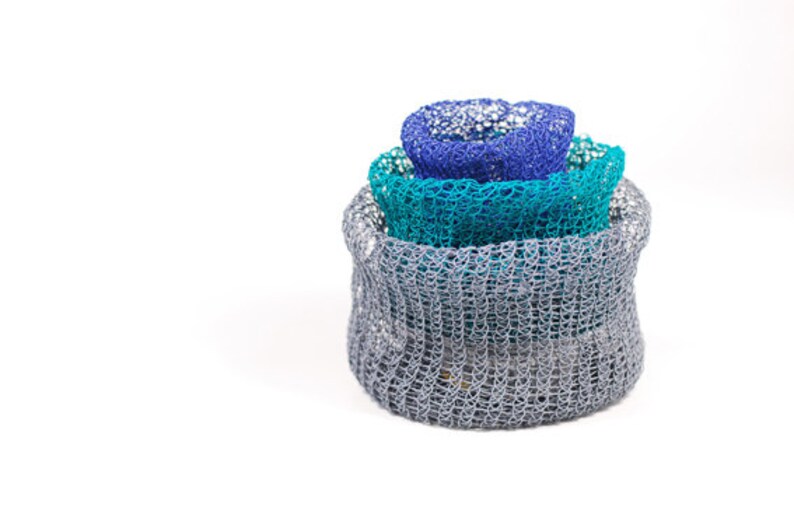 May include: Three crocheted bowls, stacked on top of each other. The top bowl is blue, the middle bowl is teal, and the bottom bowl is gray.