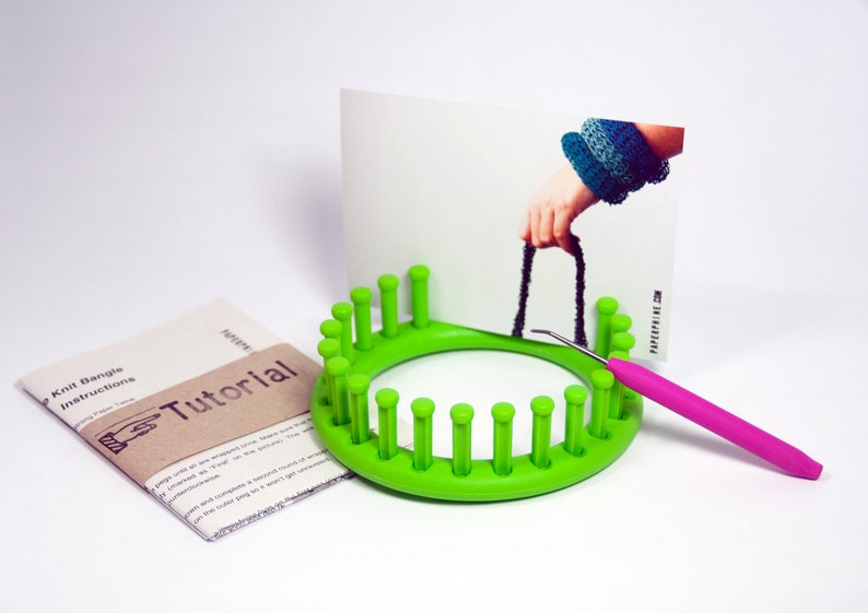 May include: Green knitting loom with 20 pegs, a pink crochet hook, and a printed instruction sheet that says "Knit Bangles Instructions Tutorial".