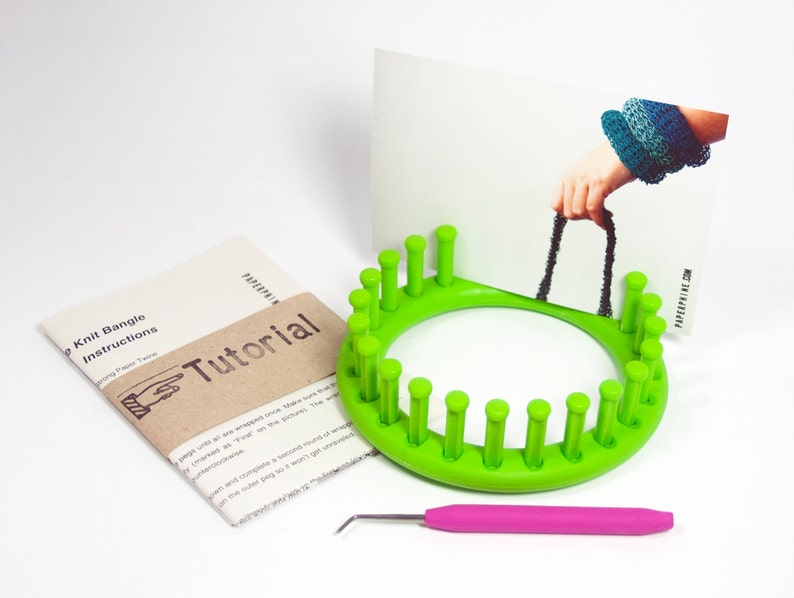 May include: Green knitting loom with 20 pegs, a pink crochet hook, and a brown paper instruction booklet with the text "Knit Bangle Instructions" and "Tutorial".