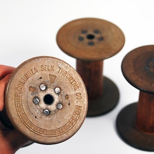 Può includere: Tre rocchetti in legno vintage con tappi terminali in metallo. I tappi in metallo sono stampati con il testo Columbia Silk Throwing Co. e presentano un foro centrale circondato da sei piccoli rivetti. I rocchetti hanno un nucleo cilindrico.