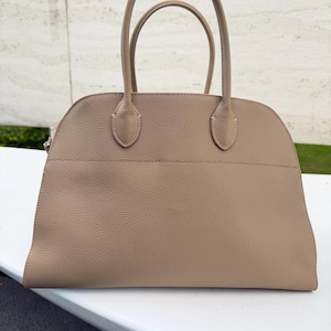 Puede incluir: Un bolso de mano de color beige con dos asas curvas y una parte superior redondeada. El bolso tiene una superficie texturizada y un dise&ntilde;o estructurado. Las asas est&aacute;n unidas al bolso con peque&ntilde;as piezas de cuero reforzadas.