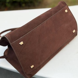 Puede incluir: Bolso de mano de ante marr&oacute;n con forma rectangular. El bolso tiene pies y herrajes de metal dorado. Las asas del bolso son visibles y el material de ante tiene una textura suave.