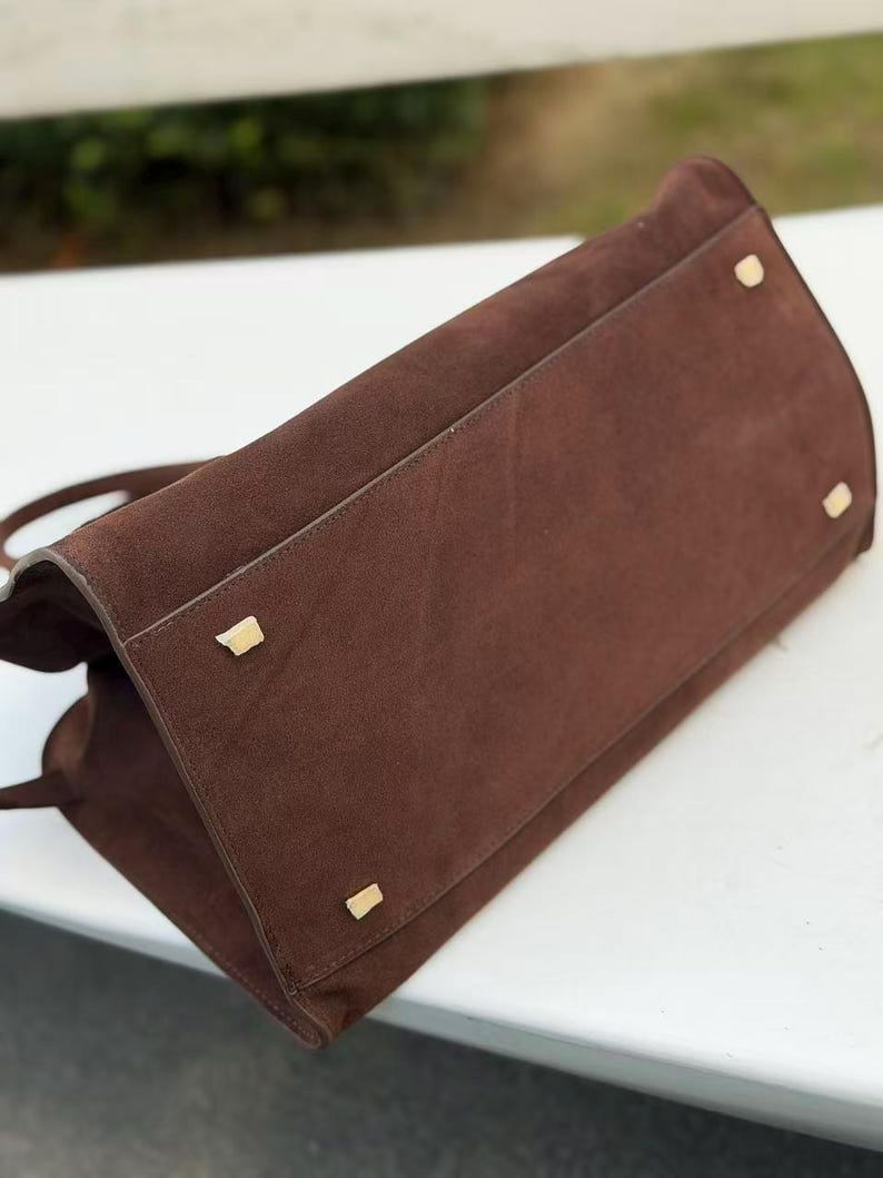 Puede incluir: Bolso de mano de ante marr&oacute;n con forma rectangular. El bolso tiene pies y herrajes de metal dorado. Las asas del bolso son visibles y el material de ante tiene una textura suave.