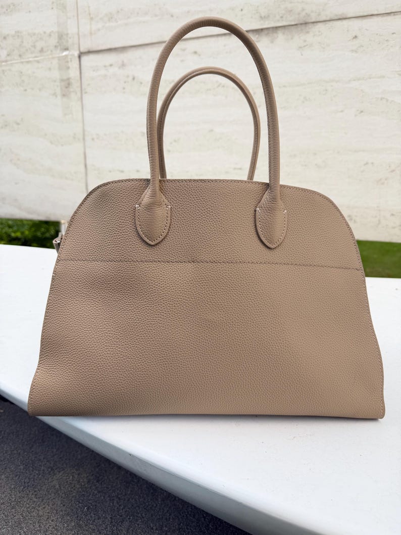 Puede incluir: Un bolso de mano de color beige con dos asas curvas y una parte superior redondeada. El bolso tiene una superficie texturizada y un dise&ntilde;o estructurado. Las asas est&aacute;n unidas al bolso con peque&ntilde;as piezas de cuero reforzadas.