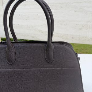 Puede incluir: Un bolso de mano de cuero marr&oacute;n oscuro con dos asas curvas. El bolso tiene una superficie texturizada y una forma estructurada. Las asas est&aacute;n unidas a la parte superior del bolso con peque&ntilde;os accesorios redondeados.