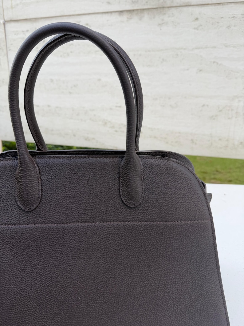 Puede incluir: Un bolso de mano de cuero marr&oacute;n oscuro con dos asas curvas. El bolso tiene una superficie texturizada y una forma estructurada. Las asas est&aacute;n unidas a la parte superior del bolso con peque&ntilde;os accesorios redondeados.