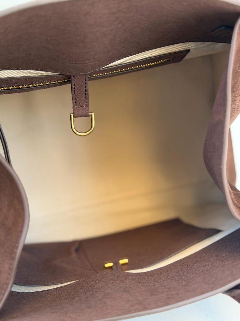 Puede incluir: Un bolso tote de ante marr&oacute;n con un interior espacioso. El bolso presenta un anillo en D y una cremallera de metal dorado, y un forro de color claro. El dise&ntilde;o del bolso es simple y elegante, adecuado para el uso diario.