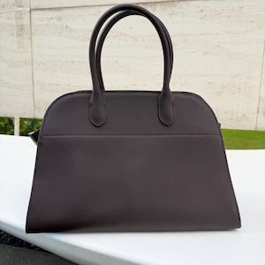 Puede incluir: Un bolso de mano marr&oacute;n oscuro con dos asas redondeadas. El bolso tiene una forma estructurada con una parte superior curva y una base rectangular. El material parece tener un acabado texturizado. El bolso est&aacute; sobre una superficie blanca.